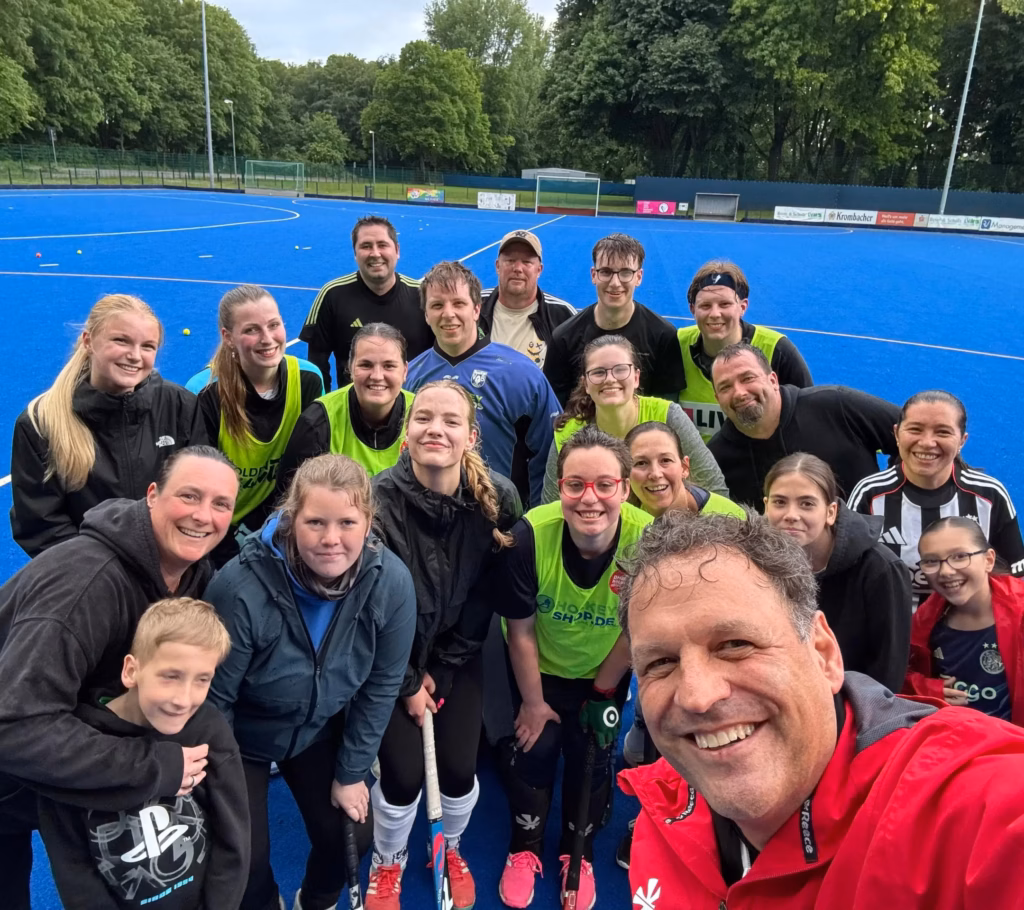Gruppe von Erwachsenen und Kindern auf einem blauen Hockeyfeld, einige tragen Sportkleidung und halten Hockeyschläger