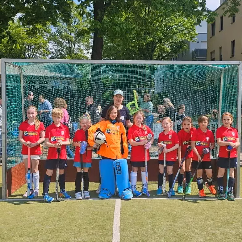 Mannschaft von Kindern in roten Trikots und ein Torwart in orangefarbenem Trikot stehen vor einem Hockeytor auf einem Spielfeld